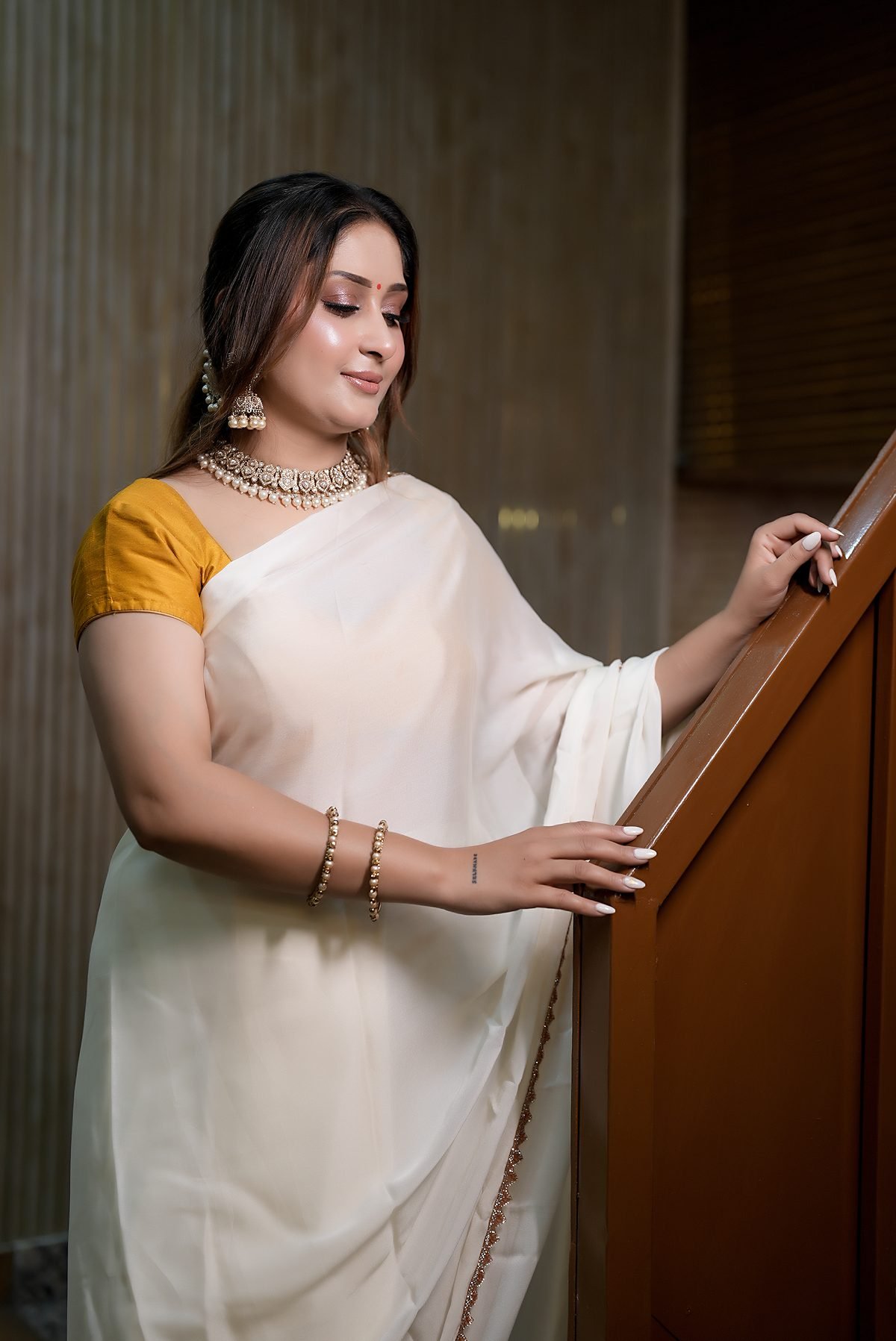 The wooden staircase up to the first floor, a woman in an ivory saree resting a hand on the rail.